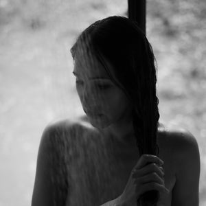 Woman standing under a gentle stream of water, photographed in soft natural light during a quiet shower moment.vvvvvv