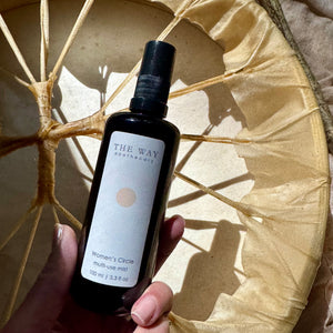 Hand holding The Way Apothecary Women’s Circle Multi-Use Mist against a natural drum surface in warm sunlight.